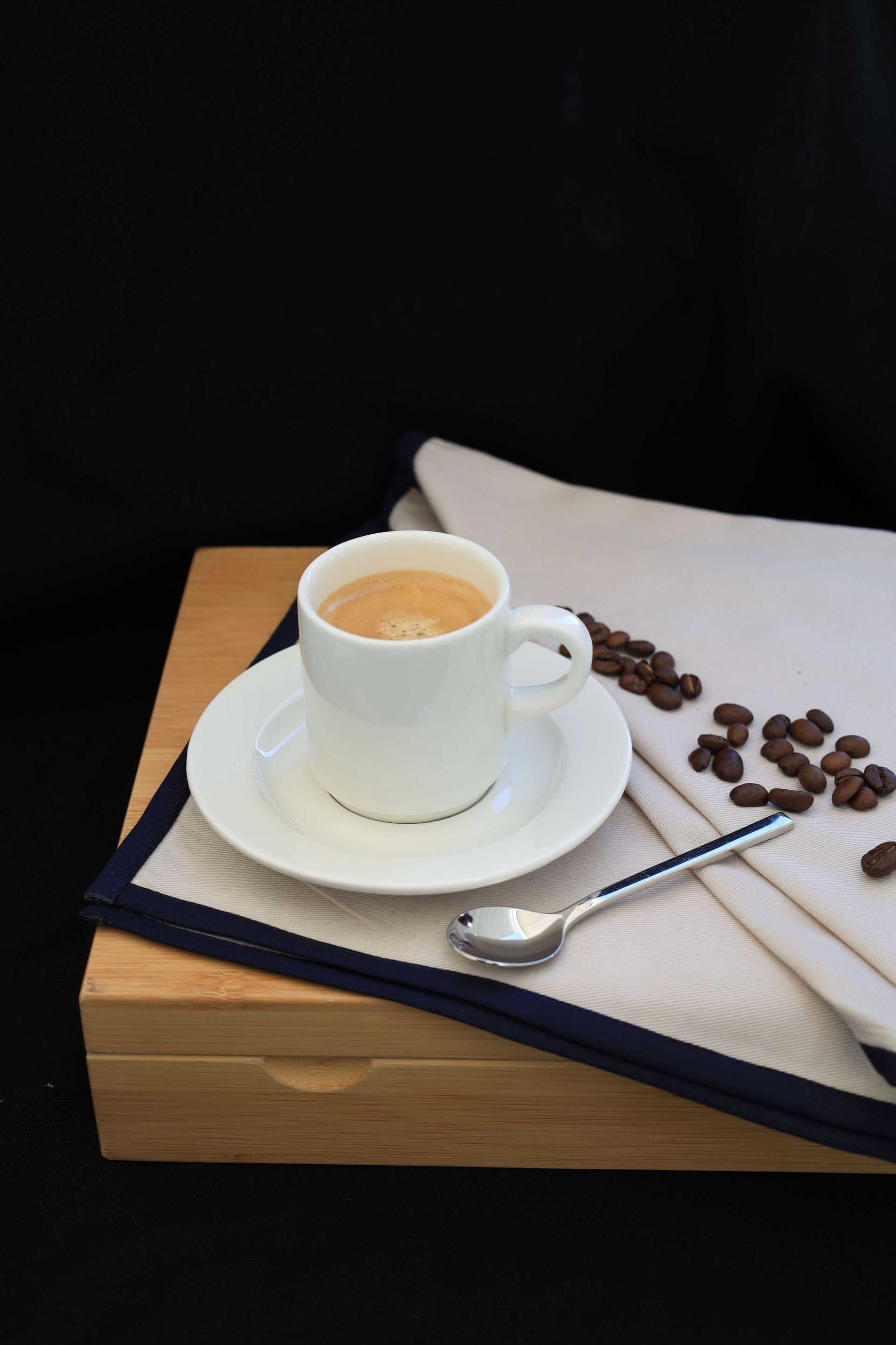 A white espresso cup on a saucer surrounded by coffee beans and a spoon on a wooden surface.
