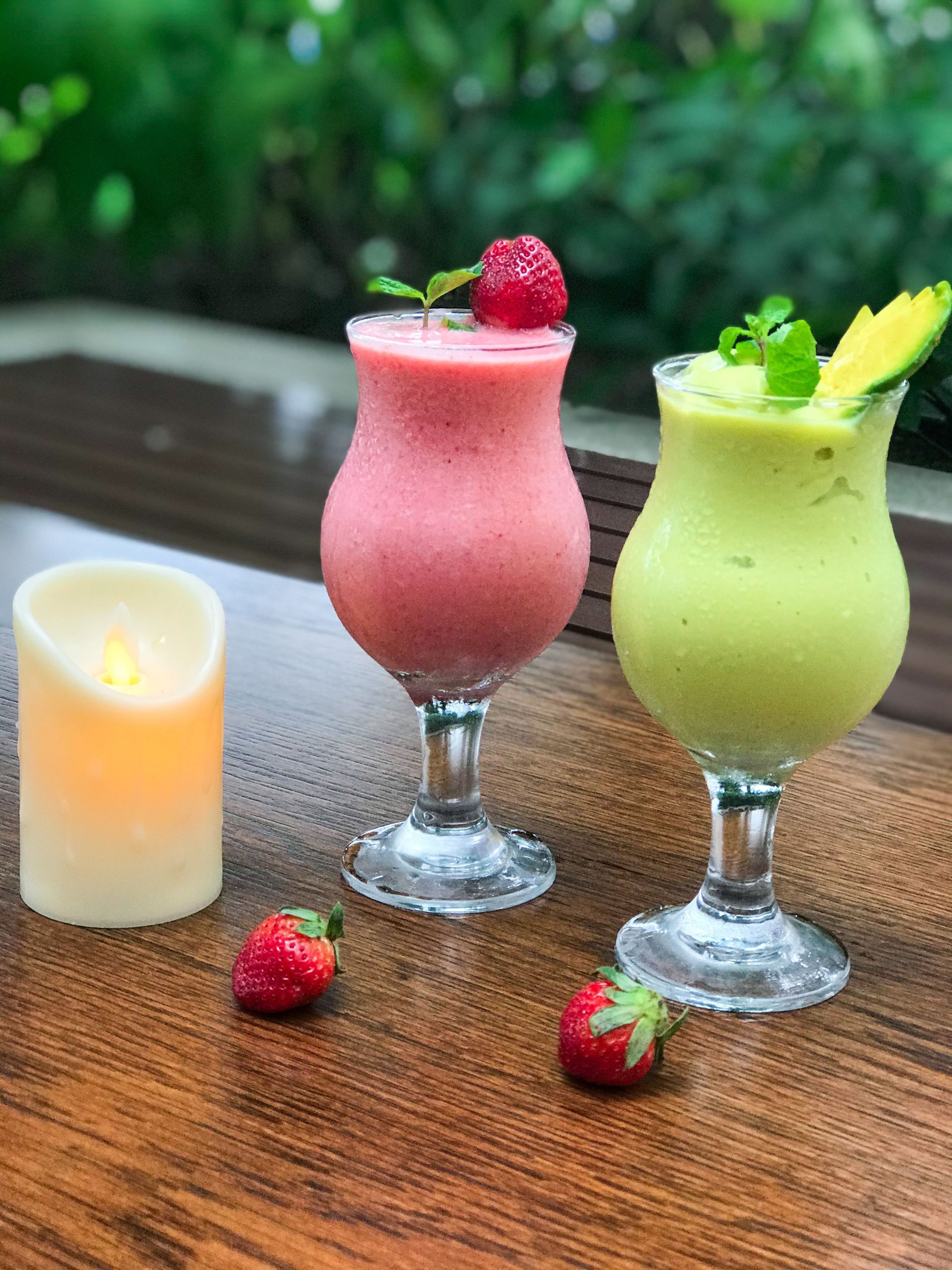 Two refreshing smoothies, one strawberry and one avocado, garnished with fresh fruits, served outdoors with a candle.